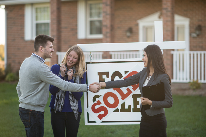 A Caucasian husband and wife are standing outdoors in front of their new house. There is a "sold" sign on the lawn. The real estate agent is shaking hands with the husband, and the wife looks extremely happy.